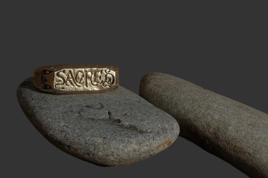 A silver engraved ring with the word 'SACRED' resting on a smooth gray stone, set against a natural background of sandy soil, larger stones, and dry twigs in dramatic, low-key lighting.